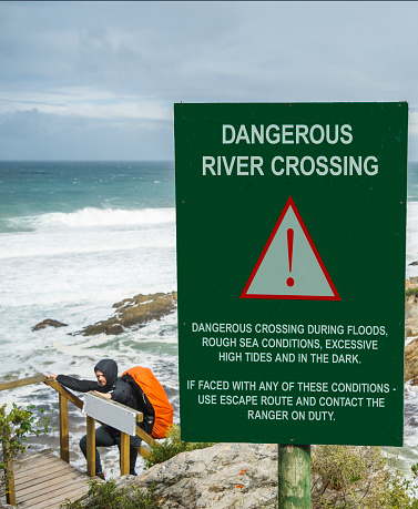 Young man in outdoor gear and carrying a backpack hiking up steps by a sign warning of a dangerous river crossing by the ocean