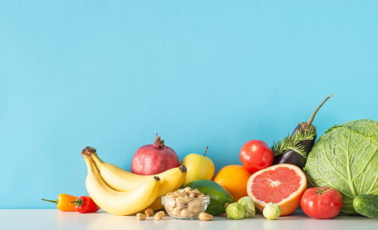 The key to vitality: Nutrient-rich diet. Side view image of a table displaying veggies (cabbage, eggplant, tomatoes, pepper) and fruits (avocado, orange, apple) on blue wall, leaving room for text