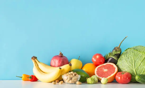 Photo of The key to vitality: Nutrient-rich diet. Side view image of a table displaying veggies (cabbage, eggplant, tomatoes, pepper) and fruits (avocado, orange, apple) on blue wall, leaving room for text