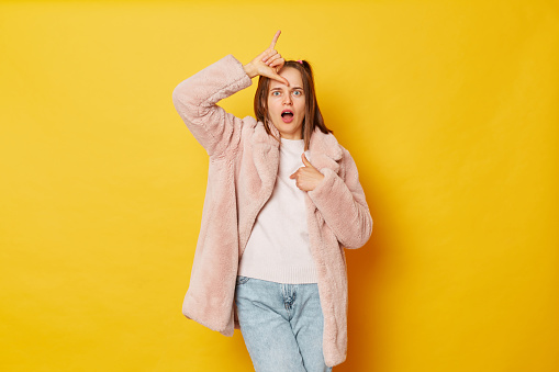 Upset disappointed woman wearing fur coat standing, showing looser gesture with hand on forehead and pointing herself looking at camera isolated over yellow background.