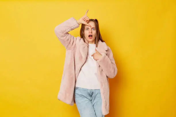 Upset disappointed woman wearing fur coat standing, showing looser gesture with hand on forehead and pointing herself looking at camera isolated over yellow background.