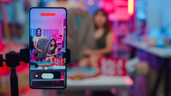 Closeup of young Asian women live streaming on phone show new collection fashion clothes to customer in shop at night. SME small business e-commerce concept.