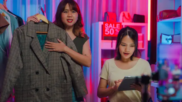 Photo of Young Asian women live streaming on phone show new collection fashion clothes to customer in shop at night. SME small business e-commerce.