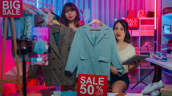 Young Asian women live streaming on phone show new collection fashion clothes to customer in shop at night. SME small business e-commerce concept.