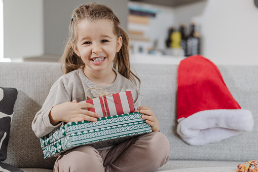 Cute little girl holding Christmas gifts in her lap while sitting on the sofa at home