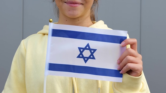 Pretty Caucasian Israeli female teenager in yellow hoodie is waving small Israeli flag, smiling at camera. Zoom out, close up, grey background, small girl is keeping flag of Israel in her hands.