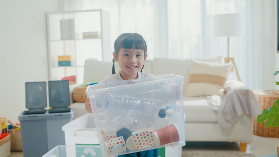 Asian girl kid holding a box full of plastic bottles to recycle happy and smiling looking at camera in living room at home. Family happy moment concept.