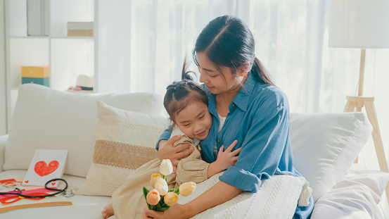 Young Asian family daughter congratulates mom and gives her flowers tulips having fun in living room during holiday celebration mothers day at home. Family happy moment concept.