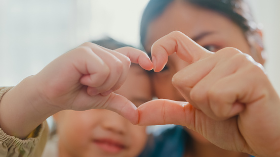 Closeup of young Asian family child making fingers heart shaped signs on sofa in living room at home. Love showing hand heart gesture, Family happy moment.