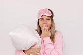 Young sleepy blonde woman in a pink pajamas and sleep mask yawning and covering her mouth with hand