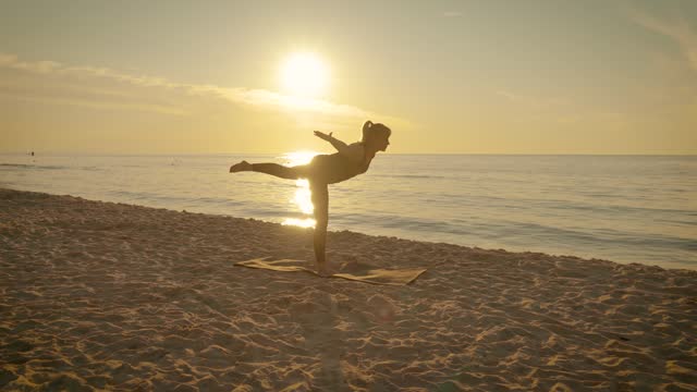 Middle years woman doing yoga performs oriental stands and balance elements at sunset beach