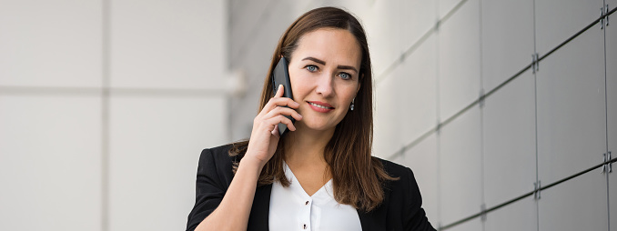 Smiling adult businesswoman dressed in formal suit stands outdoors talking on phone. Brunette woman talks on phone with colleague about deal looking at camera