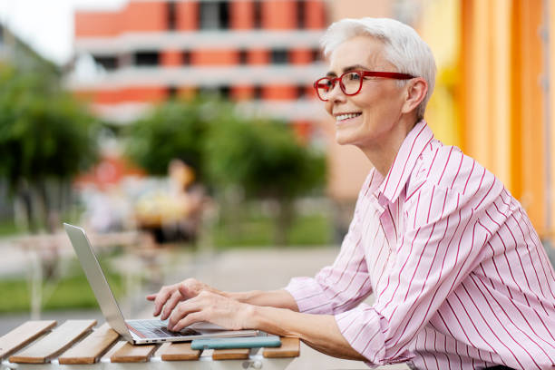 istock Positive senior woman wearing modern red glasses, using laptop outdoors, looking away, copy space 1756430747