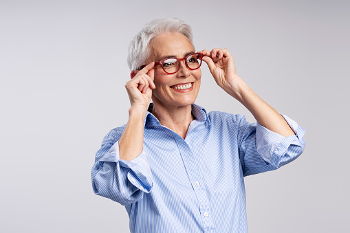 Smiling positive elderly woman wearing red glasses looking away isolated on gray background. Confident businesswoman wearing a casual shirt, health care concept