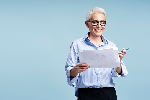 Confident smiling mature businessman wearing glasses holding business graph isolated on blue background, copy space. Beautiful teacher working with documents. Successful education concept