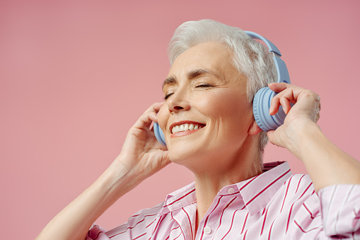 Happy senior woman with eyes closed listening music in wireless headphones, having fun isolated ob pink background. Technology, positive lifestyle concept