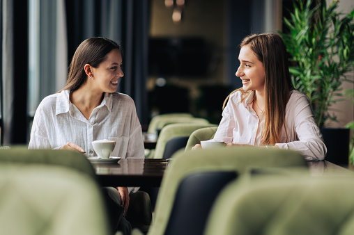 Happy female friends are sitting together in a cafe and having a coffee while chatting and smiling at each other. Girls have a coffee in a cafeteria, enjoying free time and taking a break.