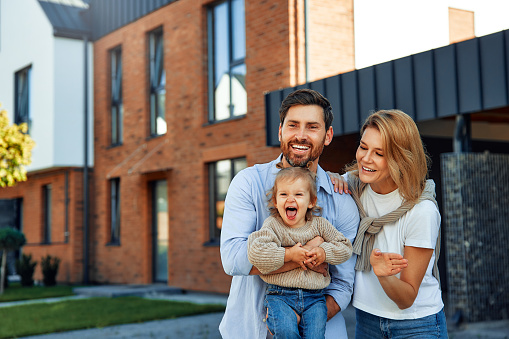 Moving of a young family to a new house. Couple with child standing in front of their newly bought house. Renting and buying a home.