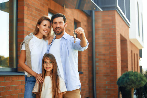 Moving, buying a home. Family shooting. A young family with their daughter standing outside their new house and showing the keys to it. Renting and buying a home. Moving concept. happy family with key of their new house stock pictures, royalty-free photos & images