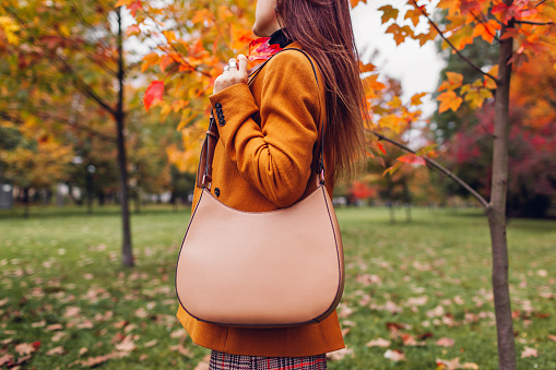 Close up of stylish woman holding handbag wearing orange blazer in autumn park among red trees picking leaves. Fall female clothes and accessories. Fashion