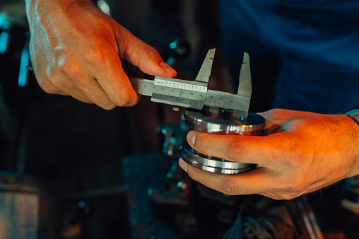 Cropped picture of a metallurgy worker measuring a bearing (metal roll cut) with a caliper. Focus on hands, work-bench setting, machine element, metal ring, milling, bored element.