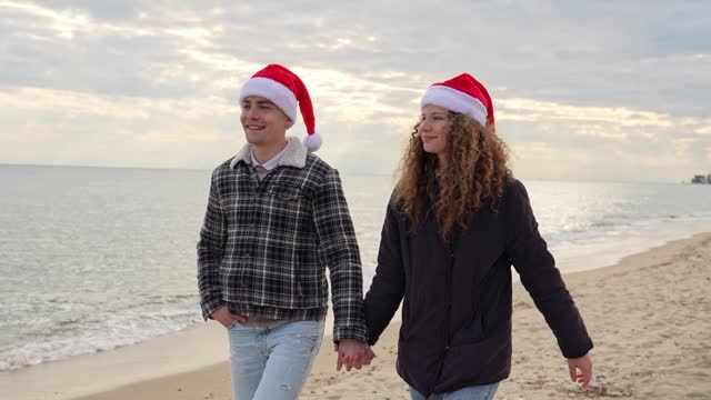 Young Couple With Santa Hats Walking Down The Beach In Greece