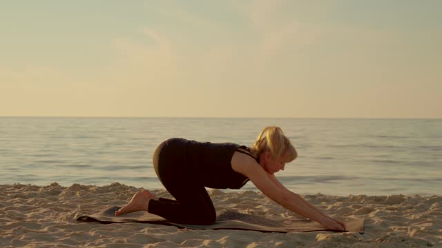 Middle years yoga woman stretching and doing flexibility exercises on fitness mat at sunset beach