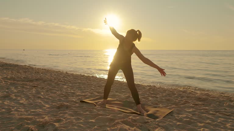 Young woman doing meditation exercise stretching sports yoga on fit mat at sunset beach