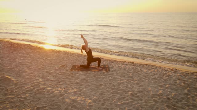 Sporty woman standing in oriental position on mat doing yoga meditation and stretching at sunset beach aerial view