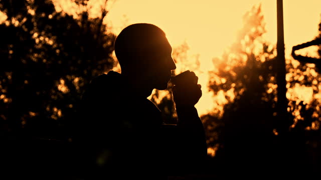 Silhouetted Man at Sunset Drinks Coffee on a Bench