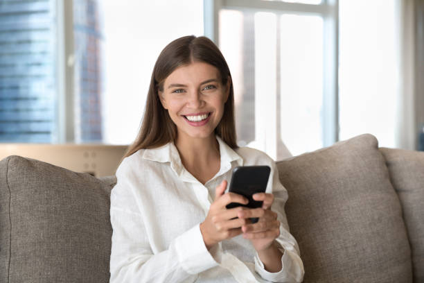 istock Woman sit on couch with cellphone smile looks at camera 1756587416
