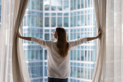 Rear view young woman standing indoors, looking out panoramic window of luxury modern apartment or hotel room opens curtains in morning, enjoying city skyscrapers view, feels happy, welcoming new day