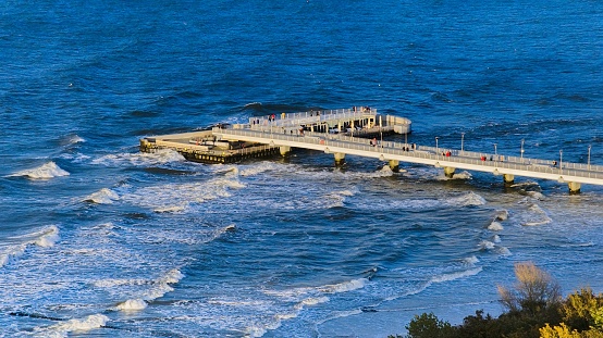 Bask in the sun-soaked serenity of Kołobrzeg's pier, as captured by this drone photo. The expansive wooden boardwalk stretches into the azure Baltic Sea, while cheerful vacationers stroll along.