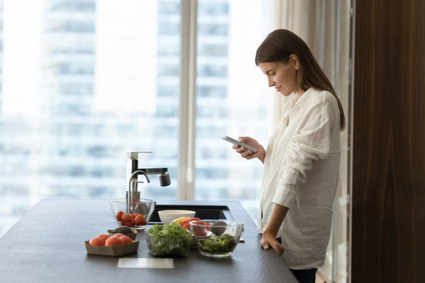 Young woman using smartphone in kitchen while cooking vegetarian salad stock photo