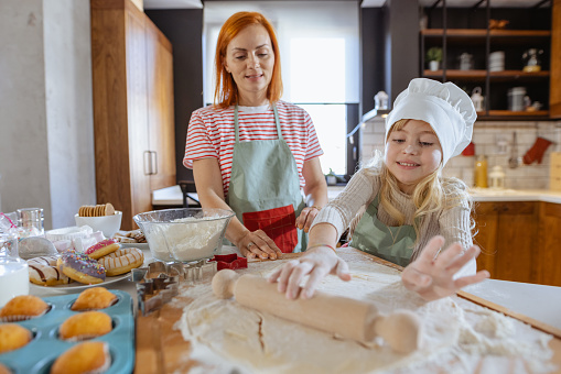 A cute girl with blond hair and her mom are wearing aprons and making cookie dough