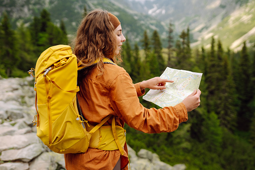 The adventure begins! Beautiful woman traveler with a yellow backpack, holds a map, explores hiking trails in the mountains. The concept of travel, vacation.