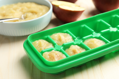 Apple puree in ice cube tray ready for freezing with fresh apple fruits on wooden table, closeup