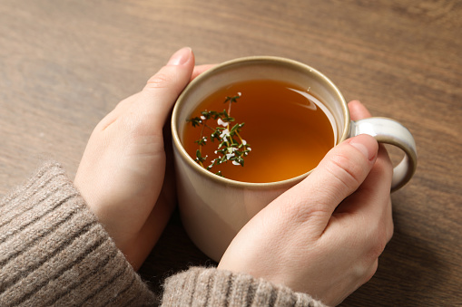 Woman holding cup of tasty herbal tea with thyme at wooden table, closeup