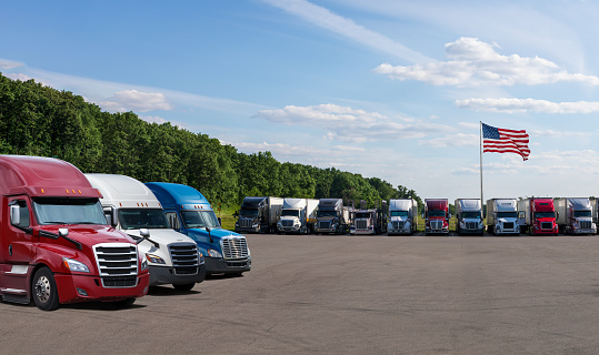 Truck stop in the USA. Different models big rigs semi trucks tractors with loaded semi trailers standing in the row on truck stop parking lot waiting for the route continuation time according to the log book.