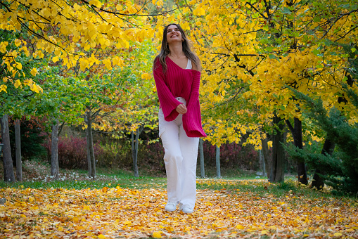 Beautiful woman walking among yellowed trees in autumn. wearing a loose pink sweater. She has sunglasses and a cell phone. Taken in daylight with a full frame camera.
