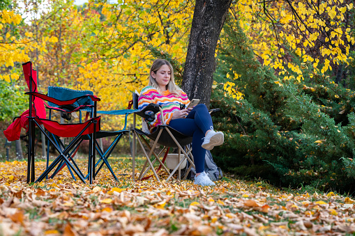 Beautiful pregnant woman sitting on camping chair among yellowed trees in autumn. wearing a loose colourful sweater. woman with blonde hair. Taken in daylight with a full frame camera.