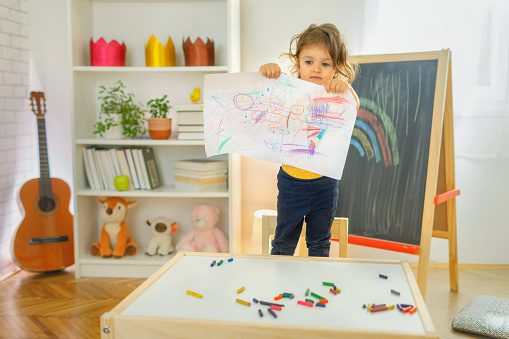 Young girl smiling and standing on a chair and holding a drawing in her hand. School education and creativity concept.