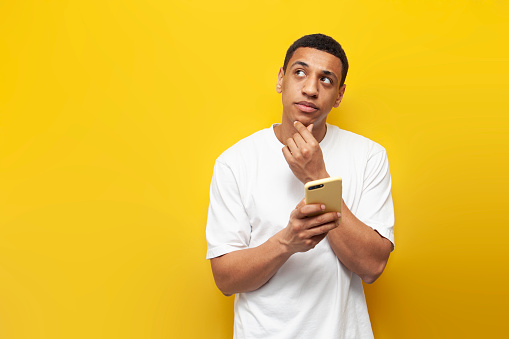 young pensive african american man in white t-shirt uses smartphone and dreams on yellow isolated background, man thinks and looks at copy space with phone