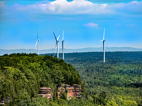 Four windmill, power turbines are erected above the the forest. There as small hill with a rock faced cliff in the foreground. The image is from around southern Ontario Canada