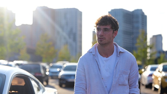 Young male in glasses walking on the parking in the city with cars on the background. Transport and lifestyle concept. Slow motion