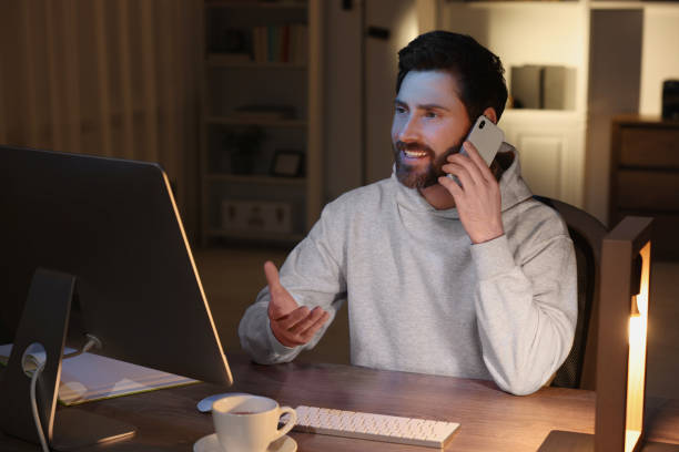 istock Home workplace. Happy man talking on smartphone while working with computer at wooden desk in room at night 1756814226