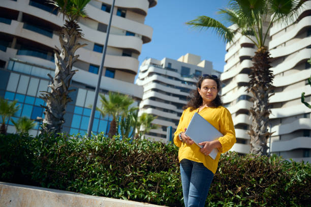 Charming freelancer woman, entrepreneur holding a laptop, smiling looking aside, standing against buildings background