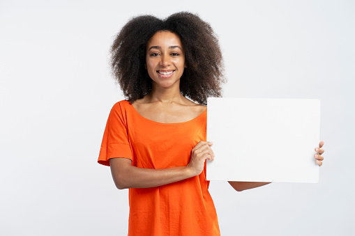 Portrait of beautiful smiling African American women wearing orange t shirt holding empty poster or banner isolated on white background, mockup. Advertisement concept