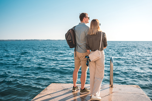 Amidst their relaxing and joyful holiday, a charming mid-adult Caucasian couple enjoys a peaceful walk in the city by the seaside, engaging in conversation, bonding, and having fun.