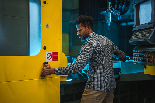 A mixed adult male production and assembly manager opening a yellow door in factory. Behind the door there is a CNC machine. The employee has his hand on the handle of the door. Behind the manager there is the control panel of the CNC machinery.
